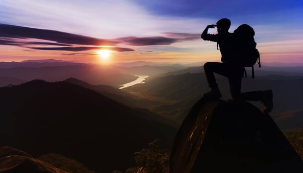 A Silhouette of a hiker on top of a mountain, shading their eyes as they look at the landscape